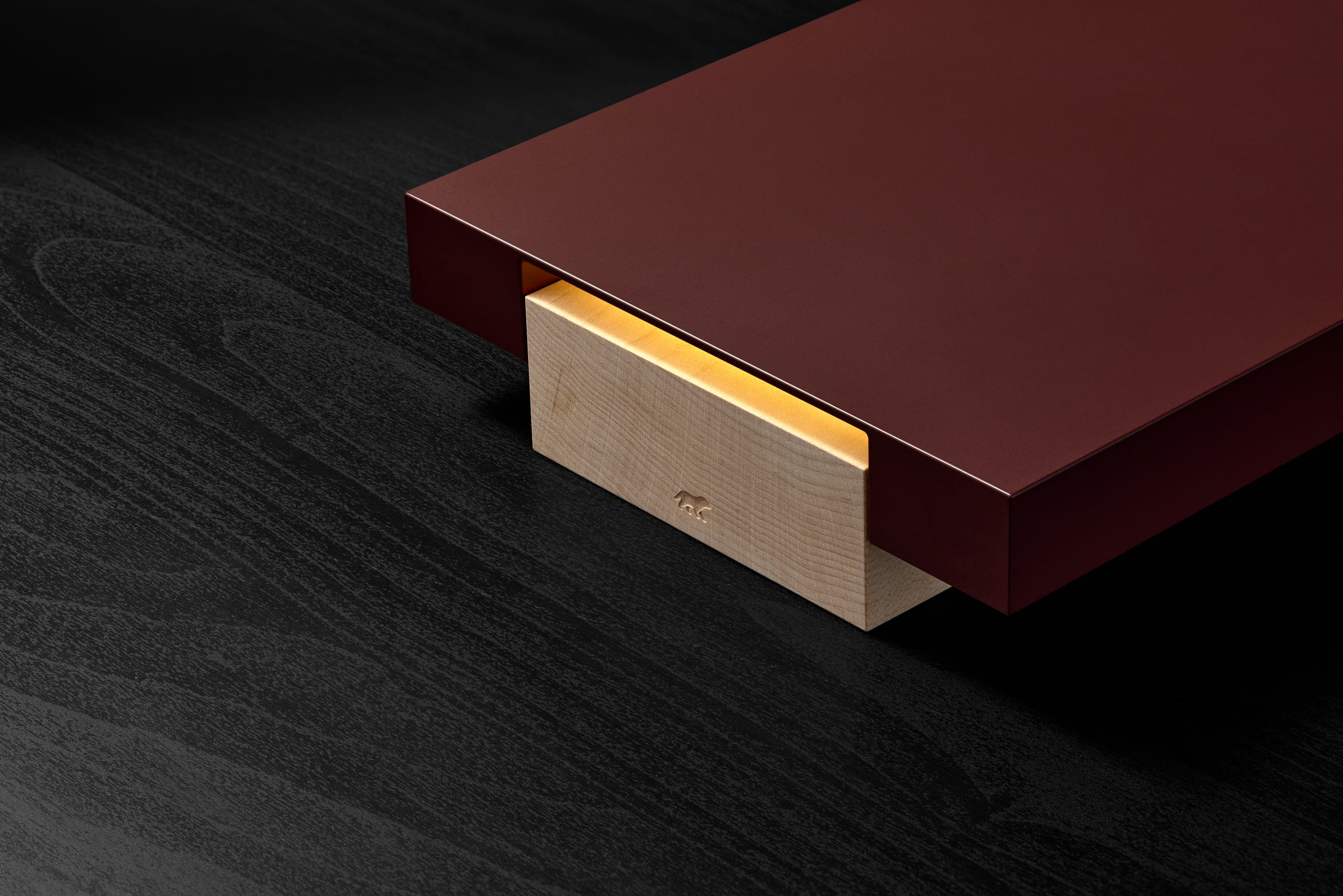 A close-up of the Vora Aegis on black-stained wood: the burgundy slab entering from the top right, its maple base lit from within.